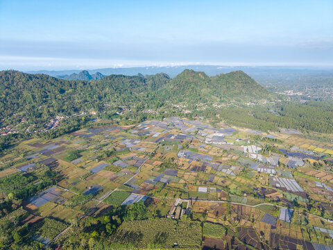 Aerial view of the agricultural fields and rural landscape of Sumberurip, featuring patchwork plots and lush green mountains in Pronojiwo, East Java, Indonesia.