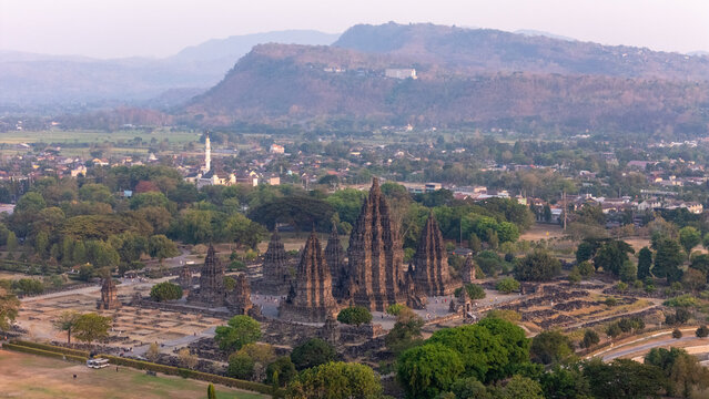 Aerial view of Prambanan Temple featuring stone spires surrounded by lush greenery, a village, and distant hills in Kecamatan Prambanan, Daerah Istimewa Yogya, Indonesia.