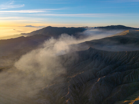Aerial view of Mount Bromo sunrise with volcanic smoke rising over the rugged caldera and surrounding mountain peaks in the golden morning light in Tosari, East Java, Indonesia.