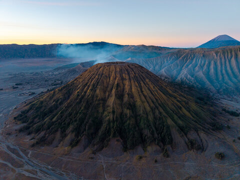 Aerial view of Gunung Batok with its distinct ridged slopes and volcanic landscape under a soft morning sky in Kecamatan Tosari, Jawa Timur, Indonesia.