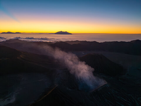 Aerial view of Mount Bromo emitting volcanic smoke at sunrise with a sea of clouds and distant mountain peaks in Tosari, East Java, Indonesia.