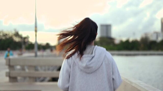 A woman is jogging along a waterfront path in sportswear, with bent arms and a steady running stride. The woman passes a railing while water and distant buildings stretch behind the pavement.