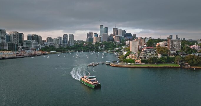 Sydney Harbour with a ferry boat navigating the water, creating a wake, with the modern architectural skyline, buildings framing the bay under a cloudy sky. Travel and explore Australia's capital city