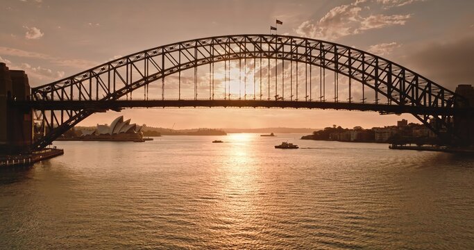 Drone flight under Sydney Harbour Bridge, the iconic Opera House stands tall in background. Creating a stunning cityscape silhouette against the glowing golden sunset reflecting on the calm waters