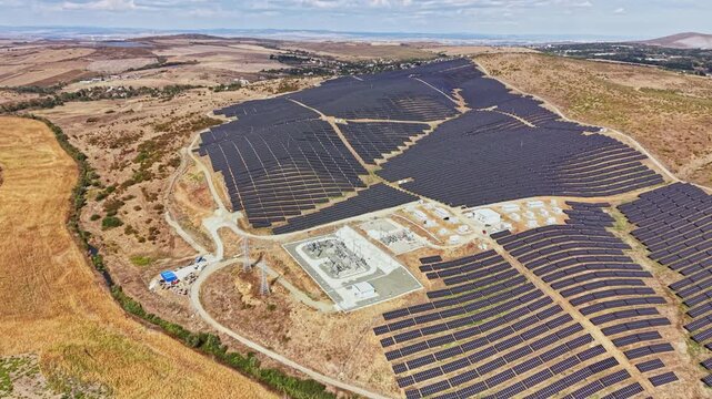 The scene shows a large solar farm spread across a dry, open landscape. Solar panels cover multiple rows in the area. The facility uses sunlight to generate energy.