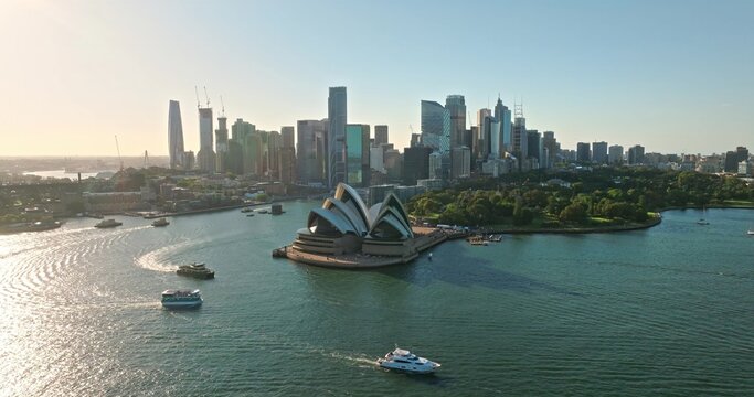 Aerial panorama of Sydney Opera House and Circular Quay waters with ferries and boats, framed by modern CBD skyline under bright blue sky on a warm, sunny day bustling with activity. Explore Australia