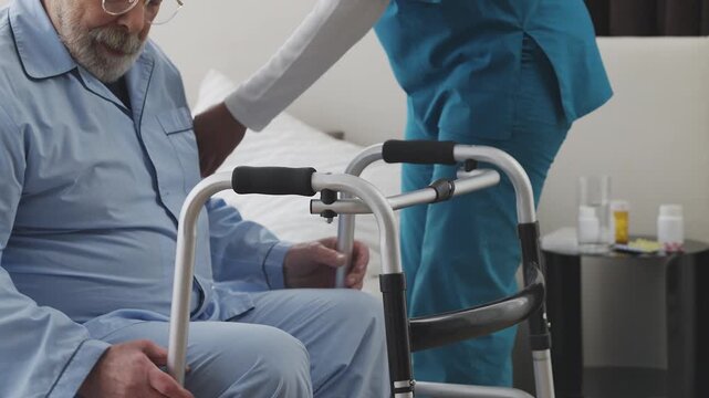 Nursing home female worker helping elderly man move with walking frame, rehab