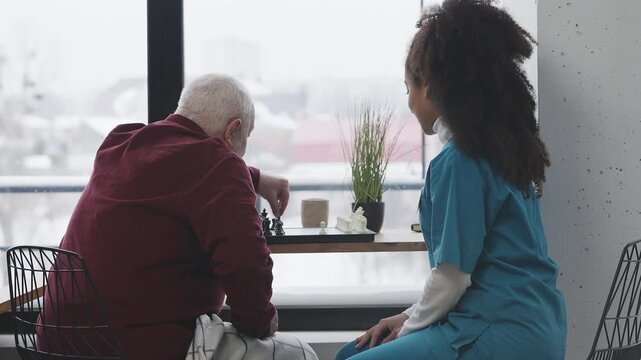 Friendly Black female carer and elderly man playing chess in nursing home ward