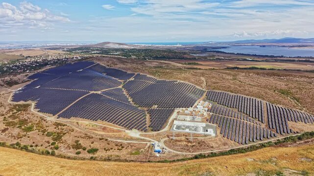 A large solar panel installation covers a hillside in a rural area. The panels are aligned in rows and surrounded by open land. The sky is bright and clear.