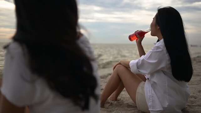 Two women sit together on a sandy shore with an ocean view and cloudy evening sky. The two women rest directly on the sand with bent or extended legs as small waves meet the beach in front of them.