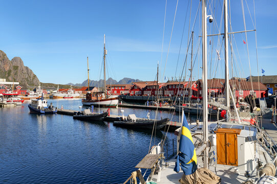 Sunny harbor of Svolvaer in the Lofoten Islands with sailboats, other vessels, red wooden houses and mountains in the background beneath a clear blue sky.