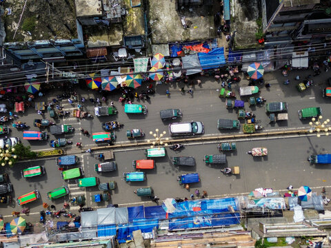 Rajshahi, Bangladesh - 26 September 2026: Aerial view of a busy street filled with colorful rickshaws, cars, and pedestrians, with vibrant market stalls and umbrellas lining the roadside.