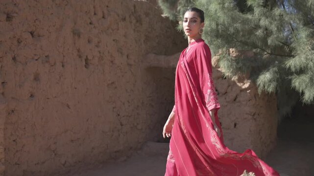 Cinematic tracking shot of an elegant woman wearing a vibrant magenta traditional dress walking past mud brick ruins in AlUla