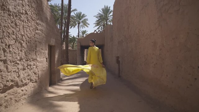 Cinematic back view tracking shot of an elegant woman in a flowing yellow dress walking between mud brick walls in AlUla palm oasis