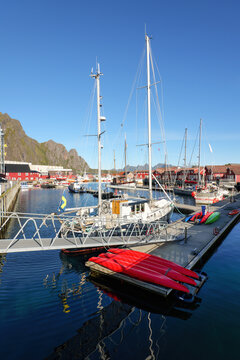 Sailboats and colorful kayaks lie in the sunny harbor of Svolvaer in the Lofoten Islands, surrounded by red wooden houses, calm water and rugged mountains.