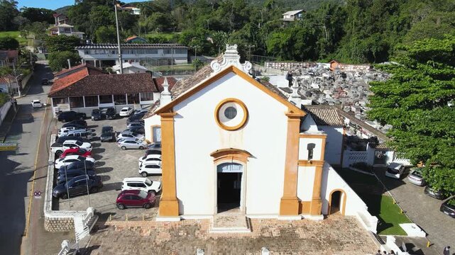 Aerial drone pull-out shot flying backward from the facade of historic Nossa Senhora das Necessidades church to reveal the square and cemetery in Santo Antonio de Lisboa, Florianopolis, Brazil.
