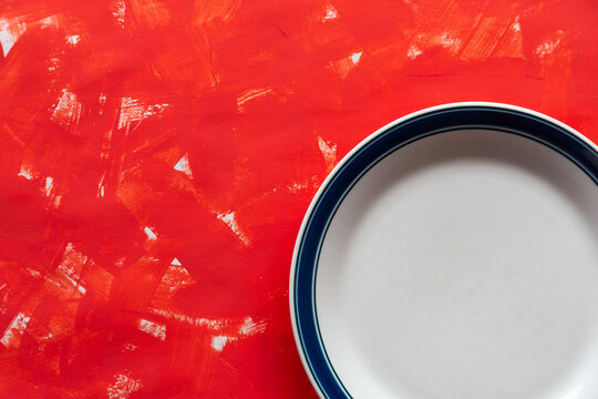 White plate sits beside vibrant, textured red surface. Bold blue rim contrasts with clean, minimalist design. Rich paint strokes create dynamic, artistic background. Ideal for culinary, design