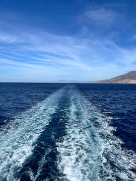 White boat wake stretching across deep blue ocean toward rugged coastal hills beneath a bright blue sky with wispy clouds, open water seascape
