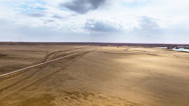 High angle shot shows a narrow asphalt road stretching across vast golden desert terrain towards distant yardang landforms under a cloudy sky.