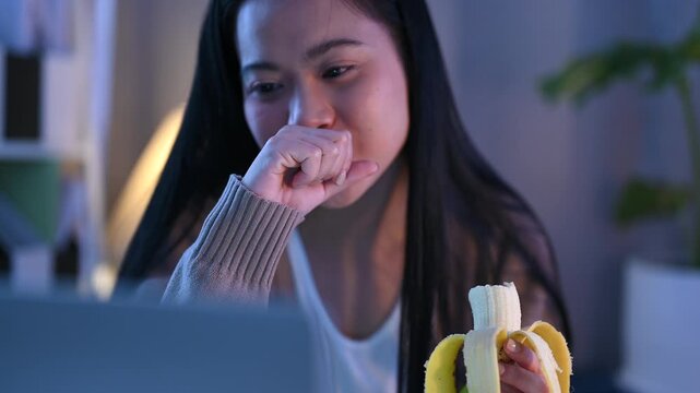 A woman eats a banana beside a laptop during night work, holding the peeled fruit near her mouth. The woman sits at a desk with papers and a lamp under cool blue light, taking a food break from work.