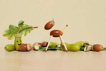 Animal figurine made of acorns and matches, green and brown acorns with oak leaves, with one painted smiling acorn, copy space for text. Autumn season © Martina