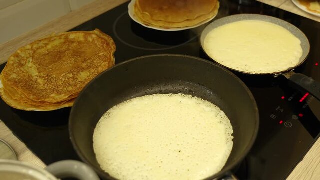 Close-up of induction stovetop with three pancakes. One cooks in a black skillet, steaming. Another rests on the counter, golden-brown. Third waits in a white bowl. Steam rises from the skillet