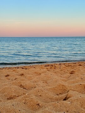 Expansive pastel sunset seascape with sandy beach foreground gentle waves distant sailboat and seagulls along shoreline under soft clouded sky and glowing horizon