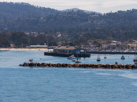 Monterey harbor pier and rocky breakwater extend into ocean waters with docked boats, distant forested coastal hills, and beachline along the California coastline.
