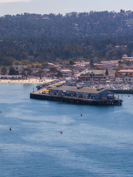 Aerial shot reveals the wooden wharf pier in Monterey, California, with kayakers paddling in the bay water, boats moored in the marina, and coastal hills visible.