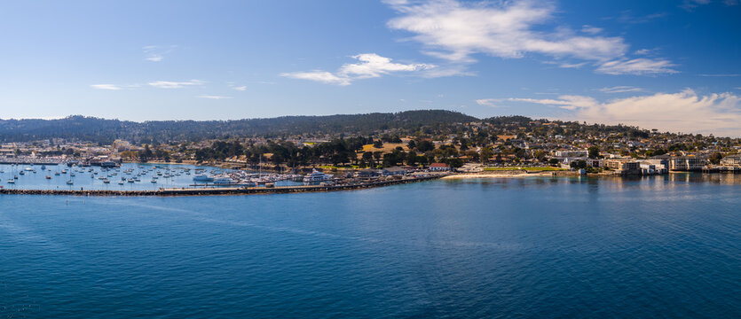 Aerial wide view displays Monterey Bay harbor with moored boats along the California coastline, featuring the marina, breakwater, coastal settlement, and distant hills.