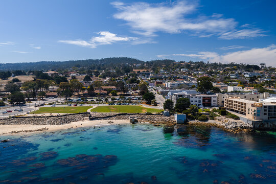 Aerial shot reveals the coastline of Monterey, California, highlighting clear turquoise ocean waters, kelp beds, a sandy beach, waterfront park, and seaside buildings.
