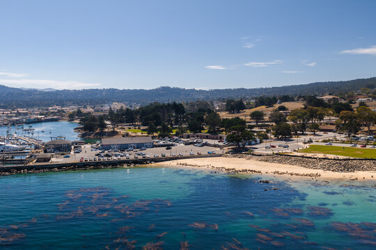 Aerial perspective of the sunny Monterey coastline in California, showing San Carlos Beach with kelp beds, the marina harbor, parking areas, and distant hills.