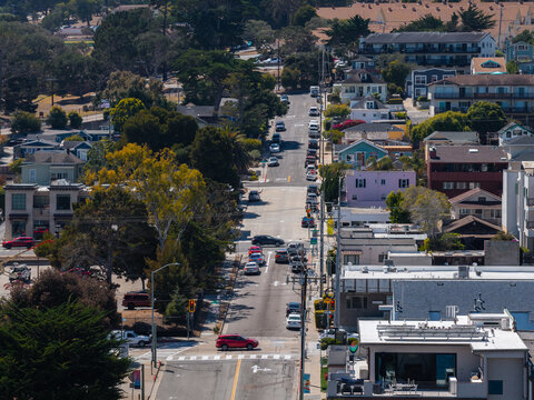 Aerial view shows a straight street lined with houses, trees, and parked cars extending towards hills under bright daylight in Monterey, California, United States.