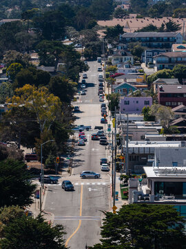 Vehicles travel along a straight, hilly residential street lined with houses, power lines, and trees in the coastal community of Monterey, California, on a bright sunny day.