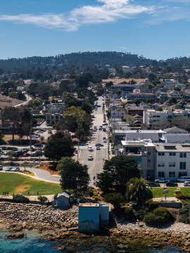 Aerial shot of a coastal street leading through the town of Monterey, California, with vehicles, houses, park areas, shoreline rocks, and forested hills under a sunny sky.