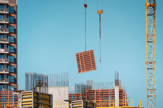 Construction site with a crane lifting a concrete formwork panel. New urban buildings rising against a clear blue sky