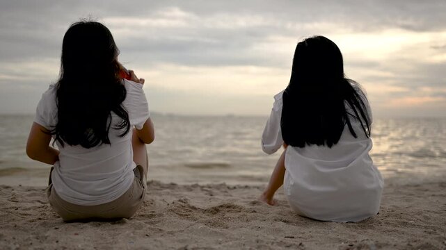 Two women sit together on a sandy shore with an ocean view and cloudy evening sky. The two women rest directly on the sand with bent or extended legs as small waves meet the beach in front of them.