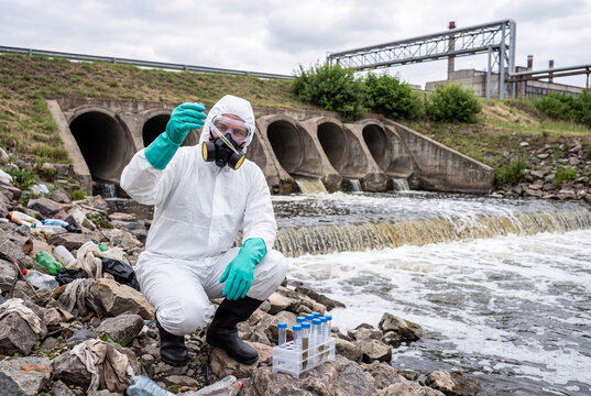 Environmental Scientist in Hazmat Suit and Respirator Collecting Water Samples for Pollution Analysis Near Industrial Drainage Discharge Culverts Quality Testing Outdoor Reservoir