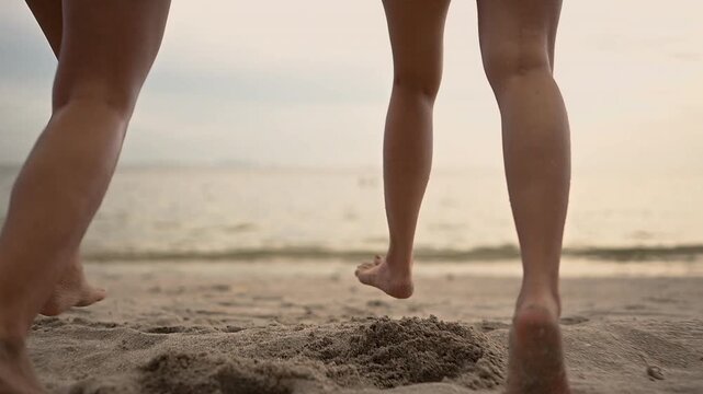 Bare feet of two women step across a sandy shore beside ocean water, leaving footprints in the textured sand. The bare feet and lower legs fill the frame while small waves wash near the shoreline.