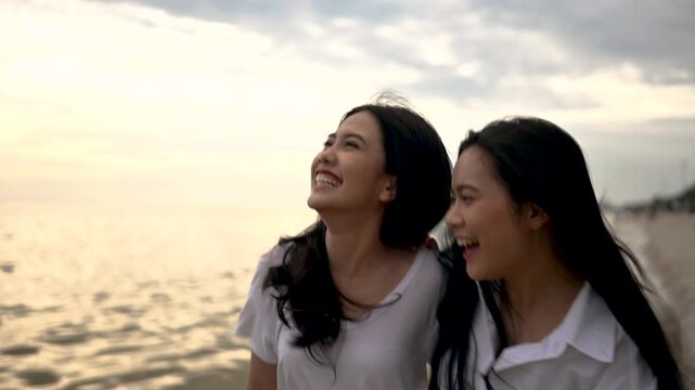 Two women walk side by side along a sandy shore in pale clothing. The two women pass ocean water under a cloudy sky, with palm trees lining the beach and open sand stretching along the coast.