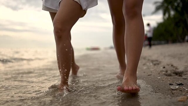 Bare feet of two women step across a sandy shore beside ocean water, leaving footprints in the textured sand. The bare feet and lower legs fill the frame while small waves wash near the shoreline.