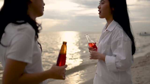 Two women hold beach drinks near the water, with glass bottles and clear cups of red liquid in their hands. The two women wear pale clothing on sand as the ocean and evening sky extend behind them.
