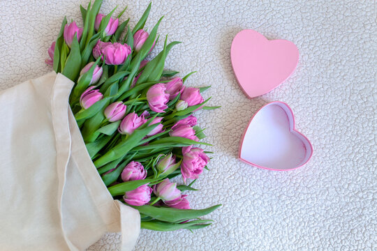 Bouquet of pink tulips in fabric bag and open heart-shaped gift box on white background. Valentine's Day, International Women's Day, anniversaries, greeting cards. Feelings of love, tenderness.