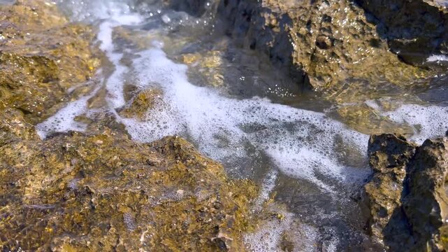 Sea Waves and White Foam Flowing Over Coastal Rocks. Close-up of sea waves and white foam flowing and swirling between coastal rocks, creating bubbles and dynamic water patterns.