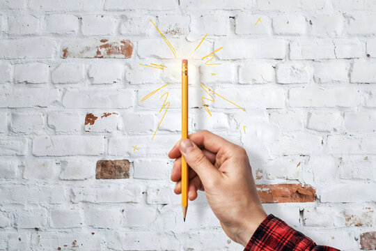 A hand holding a yellow pencil against a white brick wall with a hand-drawn yellow light bulb doodle representing a bright idea and inspiration