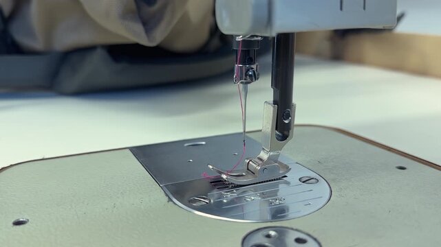 Close-up of a sewing machine needle moving down to stitch fabric on a work surface, with visible thread and machine components, showcasing the intricate process of sewing in a workshop setting