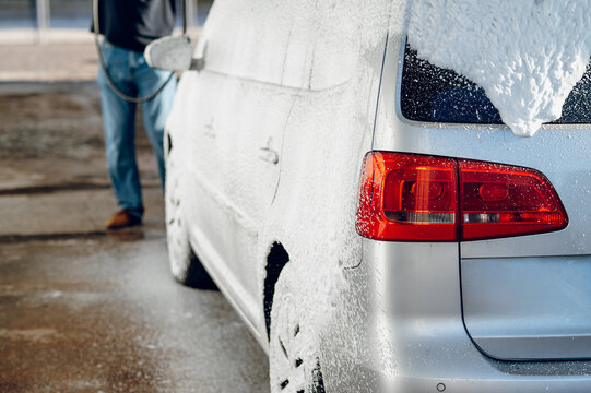 Man cleaning a silver car covered in white soap suds at an outdoor self service wash bay station