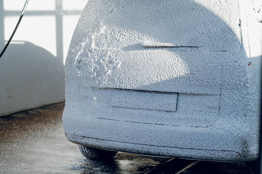 Thick white soap suds cover the rear of a vehicle during a bright daytime car wash service