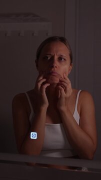 Young woman pulling down the lower part of her face in the mirror, unhappy with jowls and sagging skin during a skincare check