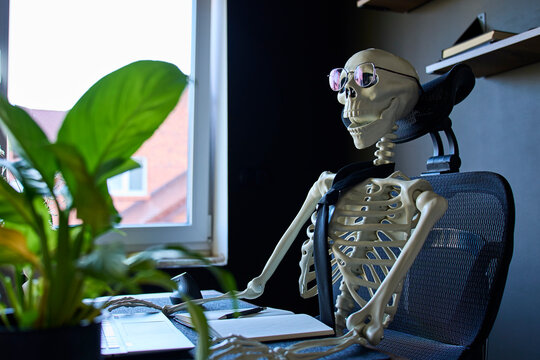 Skeleton wearing glasses and tie sitting at desk with laptop and notebook